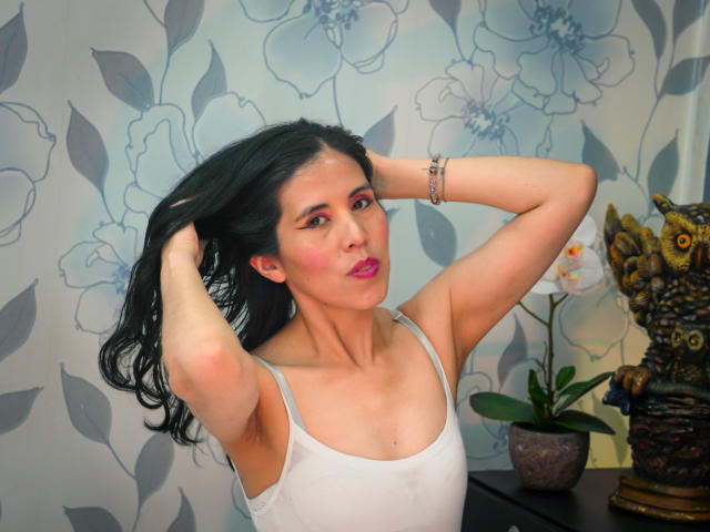 HoskLizz with black hair, pink lipstick, white tank top, arms raised, hands in hair, bracelet on wrist, looking at camera, sitting in front of owl statue and plant.