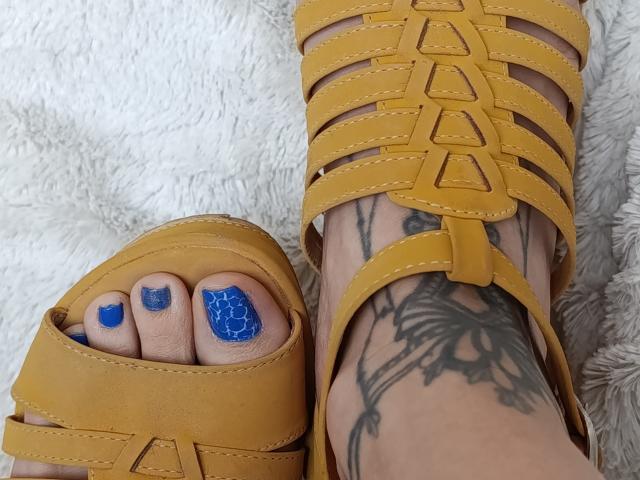 CandyParis's foot with blue glitter nail polish wearing a yellow strappy sandal, showing a tattoo of a butterfly on her ankle, next to another yellow sandal.