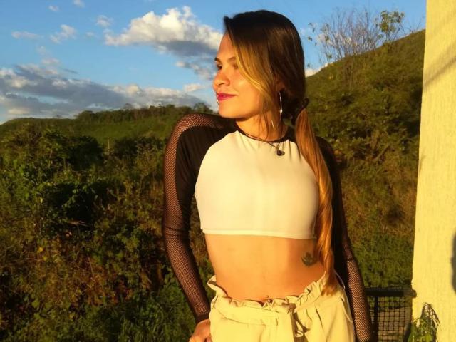 IvoryFlame with small tits, wearing a white crop top and black mesh sleeves, standing outside in sunlight, smiling, long brown ponytail, tattoo on right side, tan pants.
