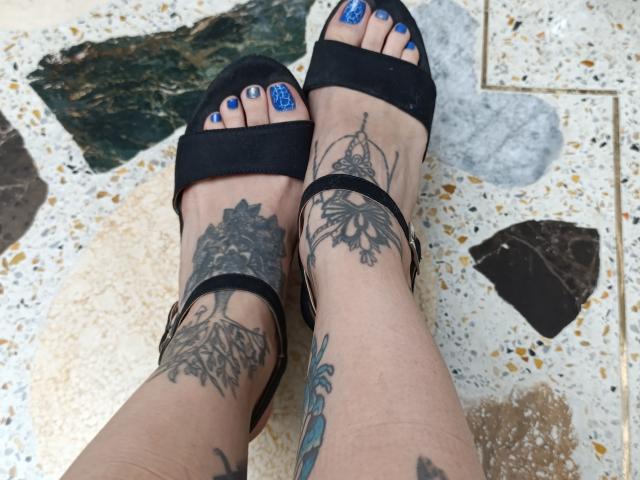 CandyParis's feet with blue nail polish, black sandals, and tattoos on her ankles and toes, showing intricate designs on her left ankle and right toe.