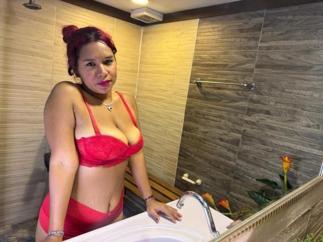 LinaBowie with red lace bra and panties, showing her big tits and curvy ass, standing in a bathroom, hands on sink, looking at camera, red hair in a bun.