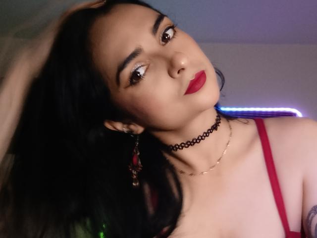 CandyParis has dark hair, red lipstick, and is wearing a black choker with red earrings and a red top, looking at the camera with a slight tilt.