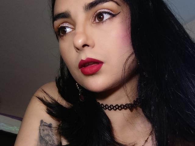 CandyParis heeft zwart haar, rode lippenstift en glinsterende oogmake-up. Ze draagt een zwarte choker en heeft een tatoeage op haar rechter schouder. Haar ogen kijken naar de zijkant..