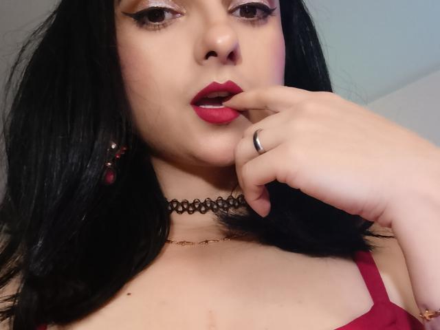 CandyParis has black hair, red lipstick, and is biting her finger with her hand near her face. She's wearing a red top and a black choker.