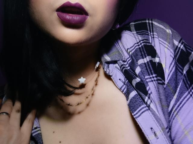 CandyParis with purple lipstick, wearing a plaid shirt, gold necklace with star, black hair, and purple background, showing her tits and hand with ring.