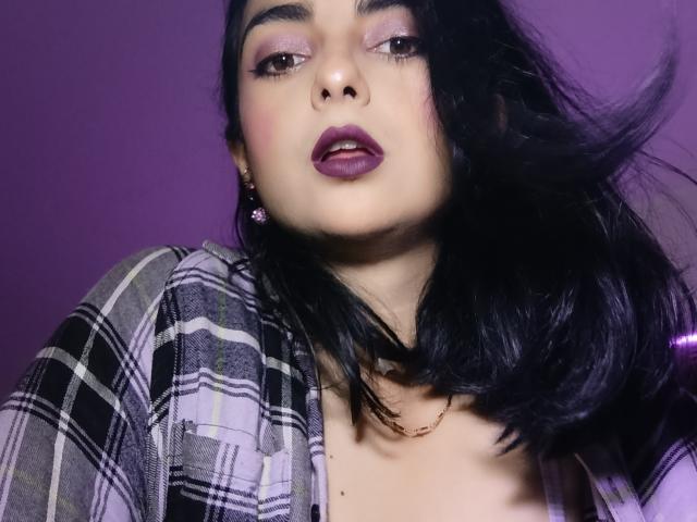 CandyParis com batom roxo escuro, cabelo preto, camisa xadrez e olhos roxos, olhando para cima com os lábios roxos ligeiramente entreabertos, com o cabelo soprando para a direita..