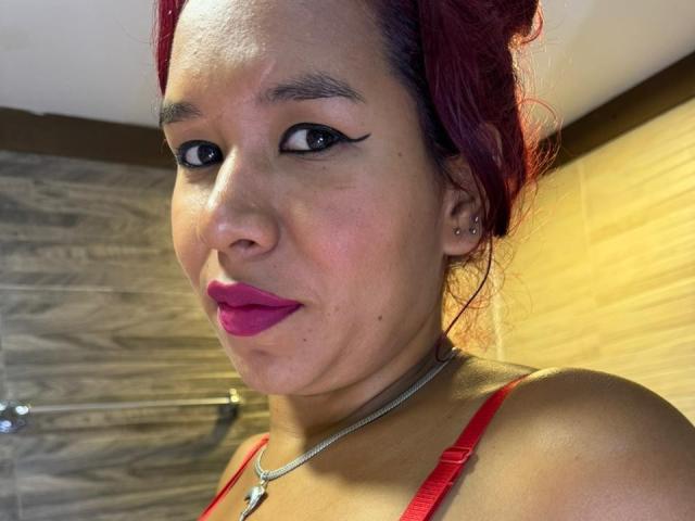 LinaBowie has red hair, black eyeliner, pink lipstick, and a silver necklace. She's wearing a red top, showing her shoulder and a small ear piercing.