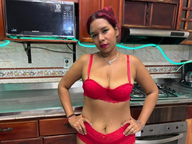 LinaBowie stands in a kitchen, wearing red lace lingerie, with visible stretch marks on her belly, hands on hips, red lipstick, and dark red hair. Boobs are large and prominent.