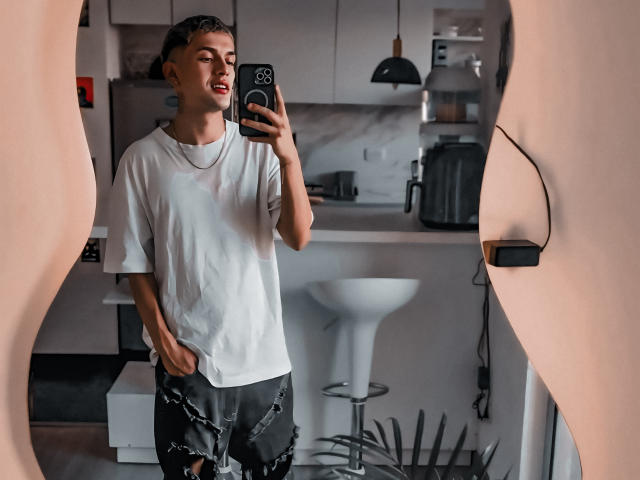 LordThoring, standing in a mirror, wearing a white tee and black, ripped pants, holding a phone, with a necklace, in a modern kitchen, looking relaxed, with a slight smile.