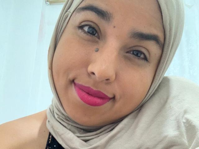 Salmahad has a tan skin tone, blue eyes, and pink lipstick. She's wearing a beige hijab and has a small mole above her right eyebrow. Her ass is not visible.