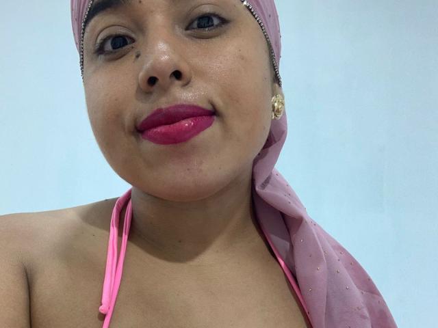 Salmahad with pink hijab, pink lipstick, and gold earrings, showing a slight smile, wearing a pink halter top, exposing her boobs. Her skin is tan, and she has small moles.