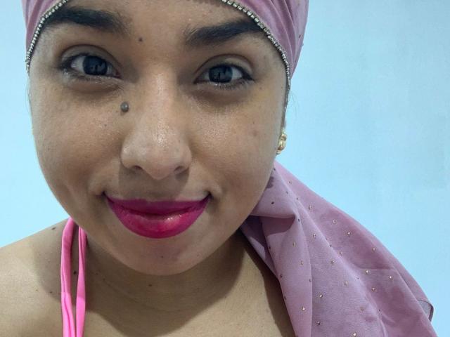 Salmahad with pink headscarf, blue eyes, and pink lipstick, smiling at the camera, showing a small beauty mark on her nose, and wearing a pink halter top.