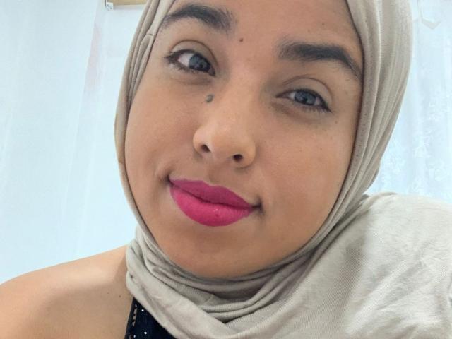 Salmahad has a light brown skin tone, blue eyes, and a small beauty mark above her right eye. She's wearing a beige hijab and bright pink lipstick.