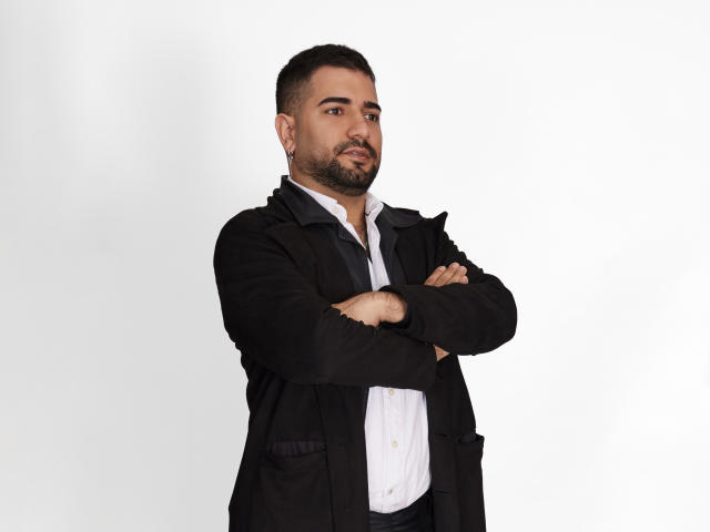 TonyLatino, a chubby dude with a beard, black jacket, white shirt, and crossed arms, looking serious, has a small nose and short hair.