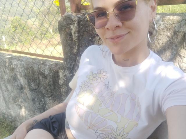 IvoryFlame with small tits, wearing a white graphic tee, black shorts, and large hoop earrings, sitting against a stone wall, wearing sunglasses, smiling, and exposing a tattoo on her arm.