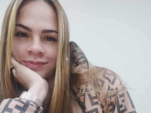 IvoryFlame, with blonde hair and brown eyes, rests her chin on her hand, smiling softly. She's wearing a geometric patterned top and a silver bracelet.