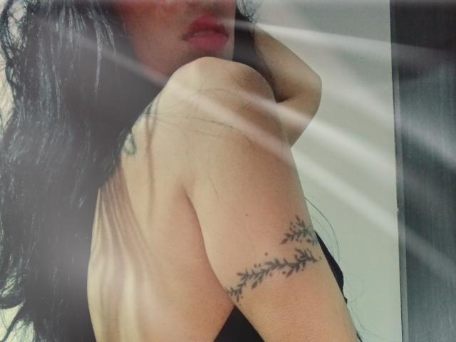 AnaisHilton's bare shoulder with a delicate branch tattoo, long black hair, and red lips, partially blurred by light rays. Her hand rests near her neck.