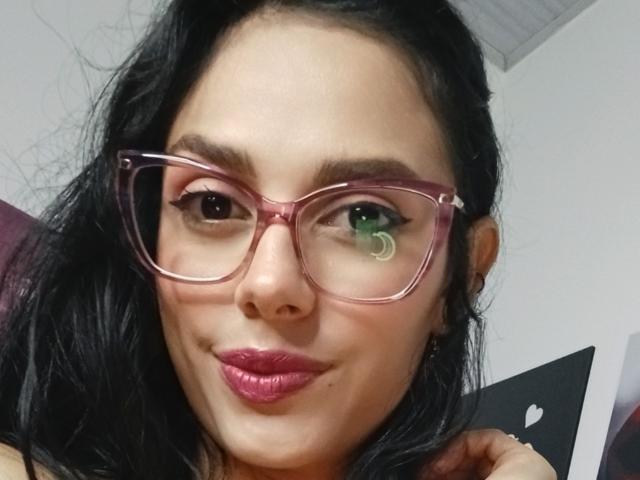 AnaisHilton with pink glasses, black wavy hair, green and brown eyes, and pink lipstick, smiling slightly, showing a small pimple on her left cheek.