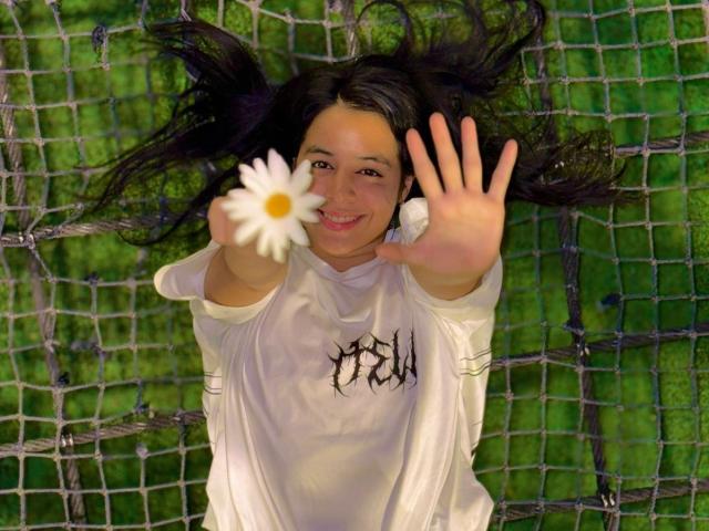 IsaBlue with long dark hair, white shirt, holding a daisy, and waving, smiling, standing in front of a net, reaching forward with her boofed hand.