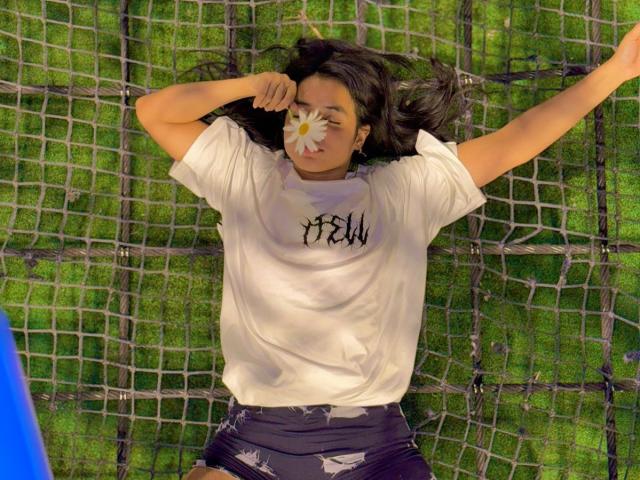 IsaBlue lying on a net, arms out, white shirt, black shorts, face covered with a flower, long dark hair, green grass background, blue object in lower left.