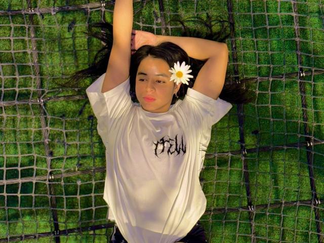 IsaBlue lying on a net with arms above head, wearing a white shirt, black pants, and a daisy in hair, green grass background, slightly smiling, looking relaxed.