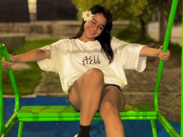 IsaBlue with a white shirt, black shorts, and a white flower in her hair, sitting on a green swing, smiling, legs crossed, wearing black socks, at night.