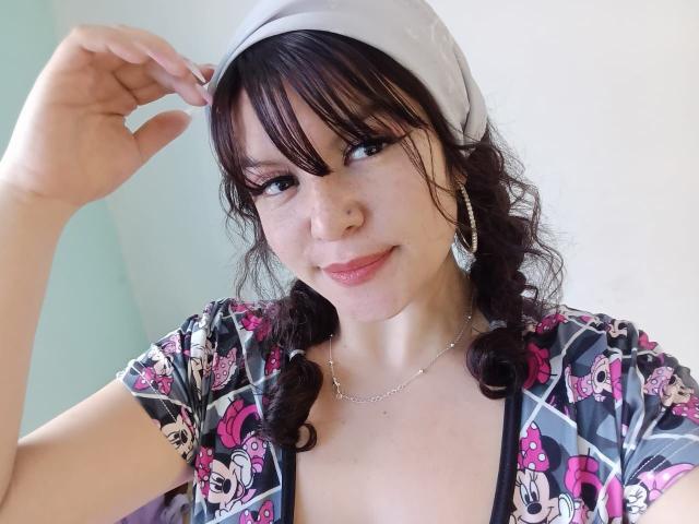 Sofinoa with dark brown curly hair, wearing a white headscarf, pink and black patterned top, smiling, touching headscarf, pink eyeliner, small nose ring, silver hoop earrings, and necklace.