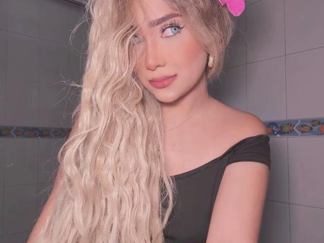 VictoriiaGrey with long, wavy blonde hair covering one eye, wearing a black off-shoulder top, pink bow, and pink lipstick, has blue eyes and a cute, sexy face.