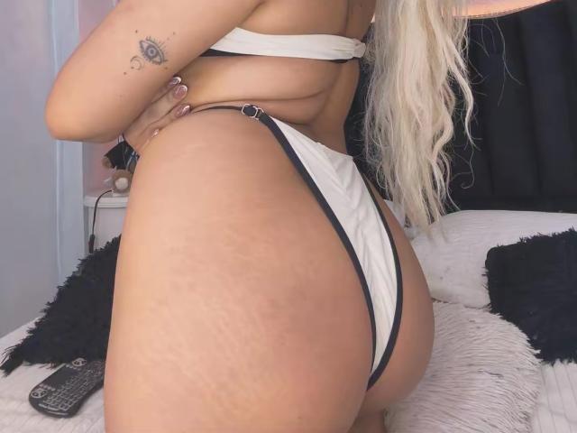 VictoriiaGrey's ass in white thong with black straps, showing stretch marks. She's wearing a white bra, long blonde hair, and has a tattoo on her right arm.