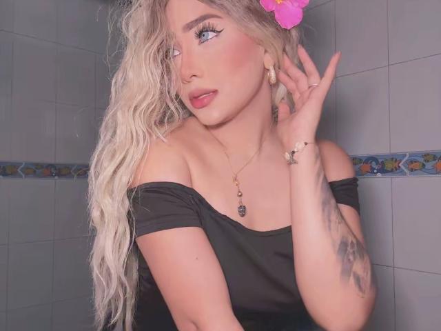 VictoriiaGrey with long, wavy blonde hair, pink flower, off-shoulder black top, tattoo on right arm, hand near face, looking to the side, wearing gold necklace.