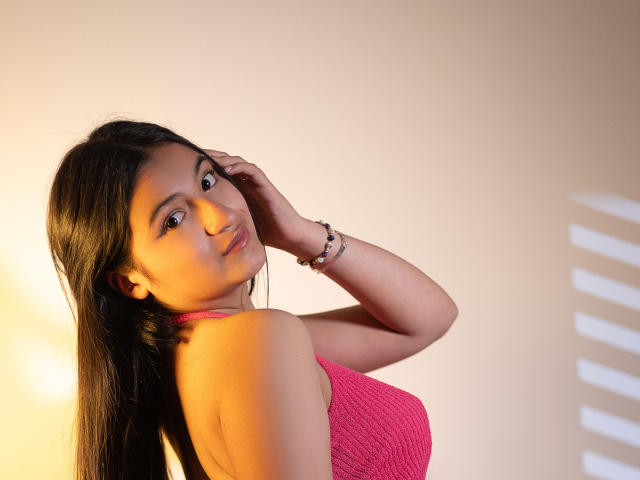 LaureneLove with long black hair, pink knit top, and bracelet, looking over shoulder, hand in hair, soft yellow light, brown skin, subtle smile, sexy, cute, attractive.