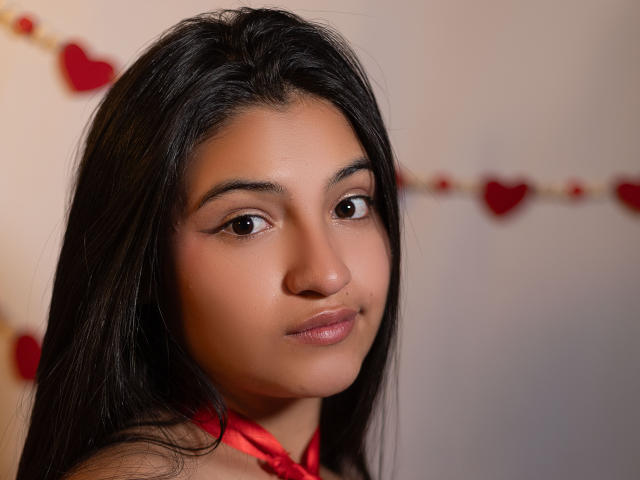 LaureneLove with brown eyes, medium brown skin, and long black hair, wearing a red halter top, has a subtle smile and a cute, sexy face.