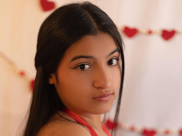 Portrait of LaureneLove with long black hair, brown eyes, and medium brown skin. She has a subtle, confident expression with slightly parted lips. She wears a red sleeveless top. The background is blurred with red heart decorations. The image has a soft, warm light. LaureneLove's makeup is minimal, focusing on her natural beauty. Her straight hair is parted in the middle. The photograph uses a close-up, slightly angled shot. The watermark "METART.com" is in the bottom right corner.