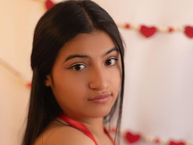 Close-up of LaureneLove, a young woman with long, straight black hair and brown eyes. She has a light brown skin tone and wears subtle, natural-looking makeup with winged eyeliner. She is looking slightly to the right with a neutral expression. LaureneLove is wearing a red halter top. The background is blurred but features red heart decorations. The lighting is soft, highlighting her facial features. The image has a warm, intimate feel.