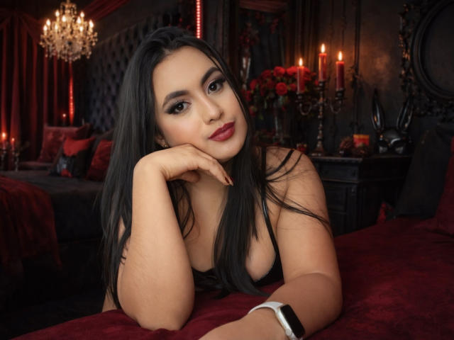ScarlettRogers with long black hair, red lipstick, and a black top, resting her chin on her hand, showing her tits, in a dark, red-lit, candle-filled room.