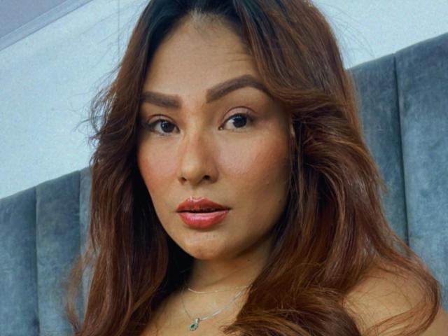 MiaaStar has wavy brown hair, dark eyebrows, and red lipstick. Her light brown skin is smooth, and she's wearing a delicate necklace with a small pendant.