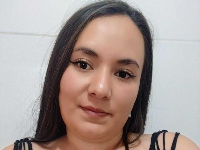 AryNoa has medium-length black hair, brown eyes, and a slight smile with subtle makeup, wearing a black top. Her skin is light, and she has small earrings.
