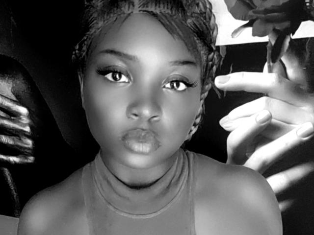 Black-and-white photo of MissAquarium with braided hair, full lips, and eyeliner, wearing a sleeveless top, hands in the background, focused on her face, looking slightly to the side.
