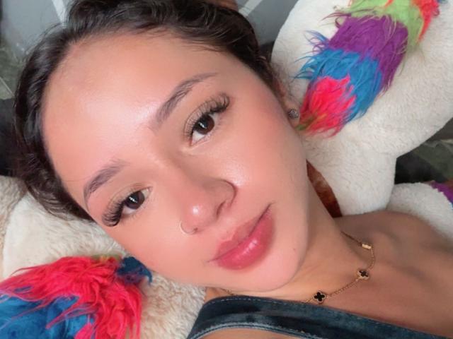 KloeThompson with a nose ring, long eyelashes, pink lips, and a gold necklace, lying on colorful stuffed animals, wearing denim. Her face is slightly flushed and she's smiling.