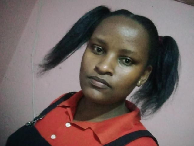 Emphi69 has dark brown skin and is looking directly at the camera with a neutral expression. She has high puffy pigtails, with black hair, and is wearing a red polo shirt with black suspenders. Her makeup includes dark brown lipstick and subtle eyeliner. She has small, gold stud earrings. The photo is slightly blurry, with a pinkish background that is out of focus. Her face is centered in the frame, and she looks slightly to the left. The lighting is even, highlighting her smooth skin and puffy pigtails.