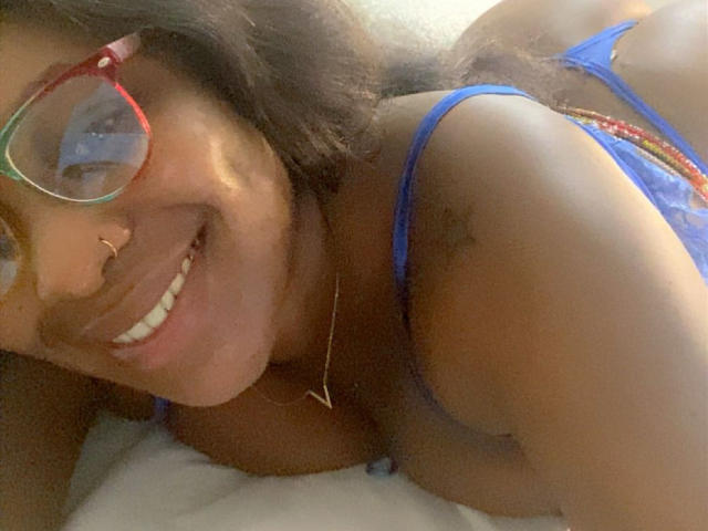 Solene69 lying on a bed, smiling at the camera with red glasses, blue lace bra, nose ring, and a gold necklace. Her brown skin and curly hair are visible.