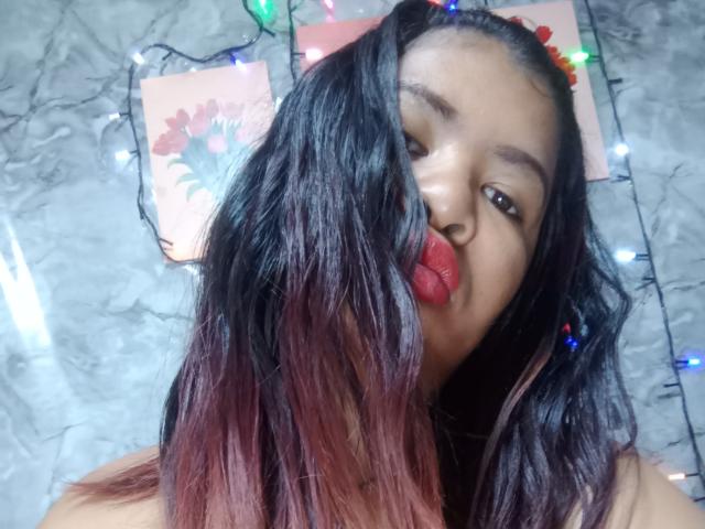 MariahSquirting69 with dark, wet-looking hair, red-tinted ends, and bright red lipstick, pouting slightly. Christmas lights and wall art in the blurry background.