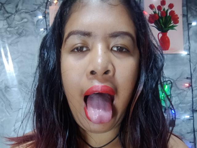 MariahSquirting69 has dark, wavy hair, red lipstick, and her tongue out, showing a pink, wet tongue. She has brown eyes and a light brown skin tone.