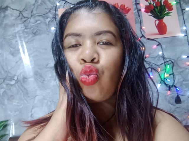 MariahSquirting69 with dark brown skin, black hair with red tips, and glossy red lipstick, pouting her plump lips, standing in front of colorful Christmas lights and flower paintings.