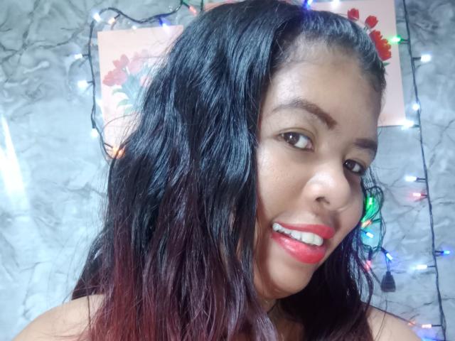 MariahSquirting69 with dark, wavy hair and red lipstick, smiling with teeth showing, wearing colorful Christmas lights in background, gray marble wall behind her.