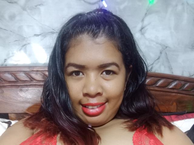 MariahSquirting69 has dark brown hair, red lipstick, and is wearing a red lace top, smiling with her teeth slightly visible. She's a curvy Asian woman with a cute face.