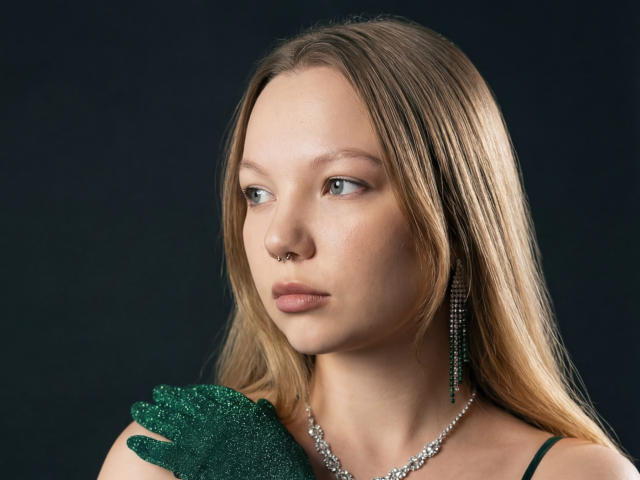MariaRobinson with green glitter glove, nose ring, and long earrings, looking off to the side. Blonde hair, light skin, wearing a necklace, dark background.