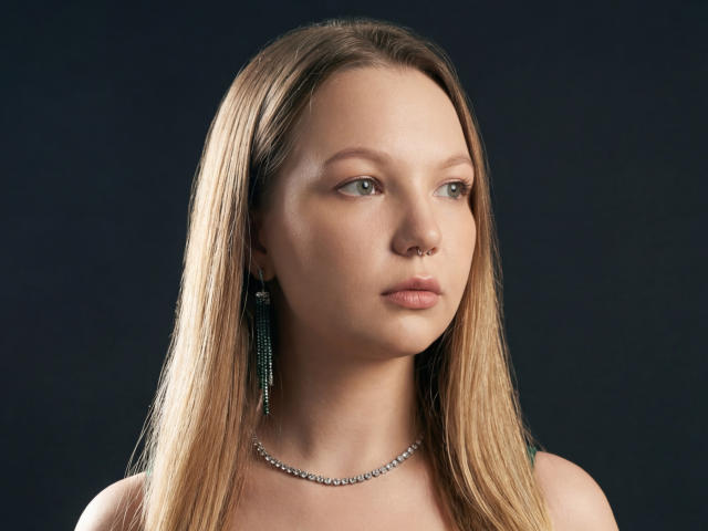 MariaRobinson has straight, medium-brown hair falling past her shoulders. She has light skin, blue-green eyes, and a small nose ring in her right nostril. She wears long, green bead earrings and a silver chain necklace. Her expression is neutral, with slightly parted lips. The image is a high-quality, close-up portrait with a dark background. The lighting highlights her smooth skin and subtle makeup. Her hair is parted in the middle, and the light source is from the front, creating a soft glow on her face.