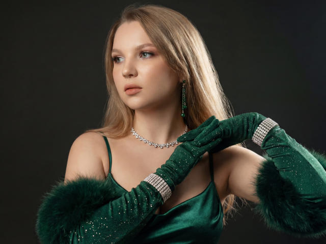 MariaRobinson has light blonde hair and blue eyes, looking to the side with a neutral expression. She wears a green satin dress with spaghetti straps, green feathered gloves, and a silver necklace. Her right hand touches her left glove, which has a jeweled cuff. She has a green earring in her left ear. The green and silver contrast with her fair skin. The lighting highlights her facial features and the texture of the dress and gloves. The image is a studio portrait.
