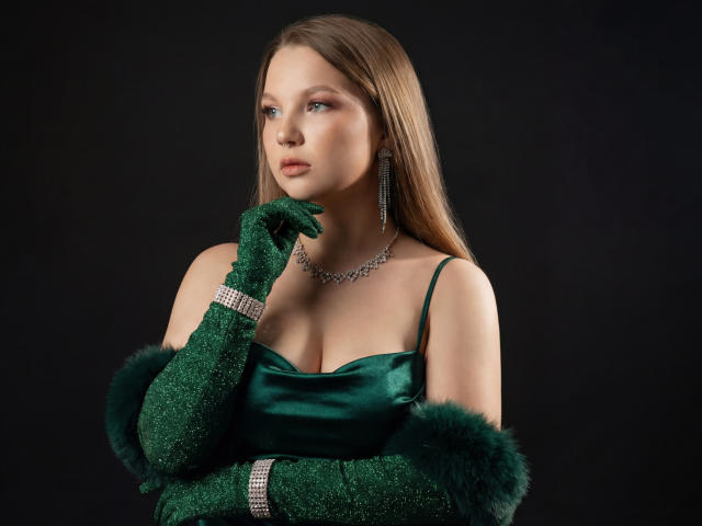 MariaRobinson with green glitter gloves, green satin dress, fur cuffs, diamond necklace, and earrings, looking off to the side with a serious expression. Boobs visible under dress.